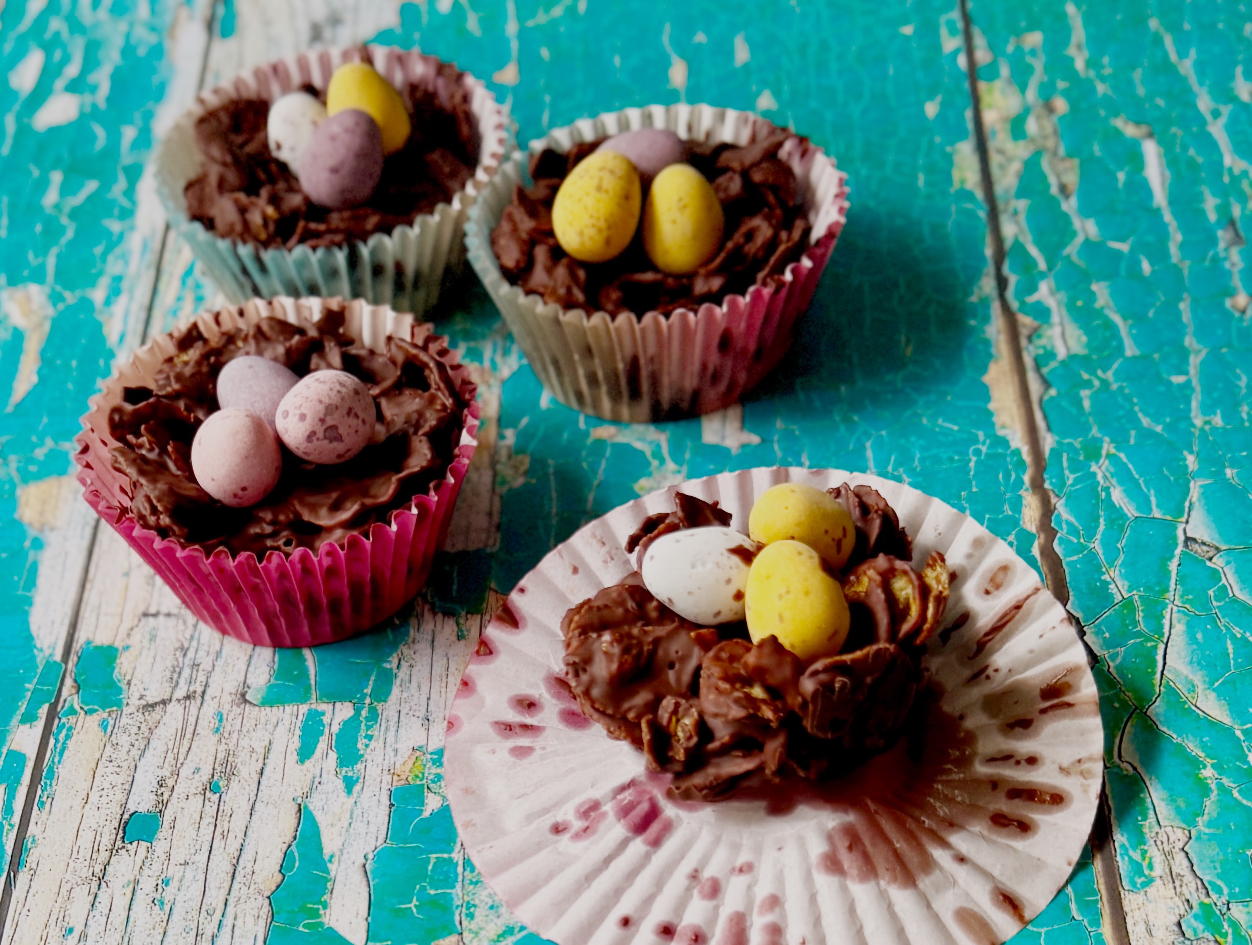 Close-up of a partly eaten Easter cornflake cake in a paper case, revealing the crisp chocolate cornflake centre with more cakes behind.